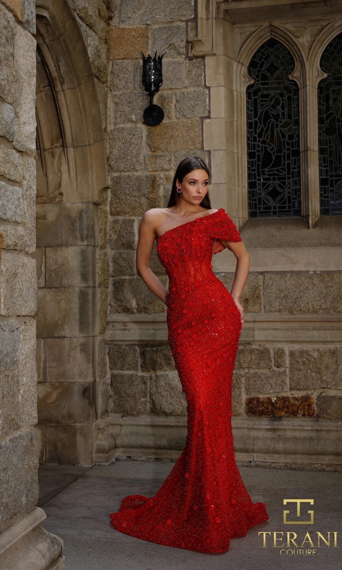 A woman in a fitted, off-shoulder red Long Formal Dress: Terani 261GL6756 stands before a stone wall and arched window, with the